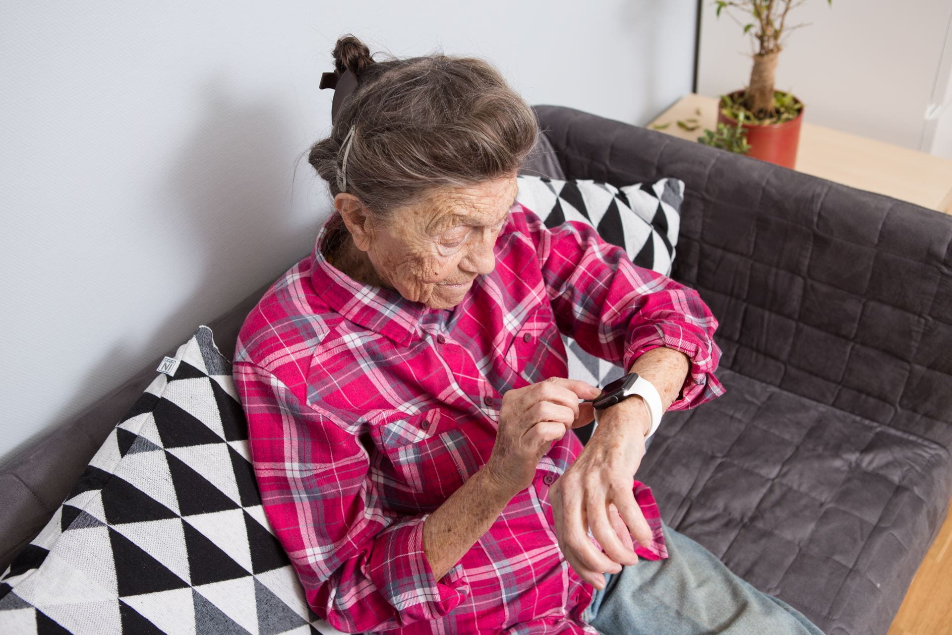 A very old senior Caucasian grandmother with gray hair and deep wrinkles sits at home on the couch in jeans and a red plaid shirt and uses a smart watch on her wrist. 